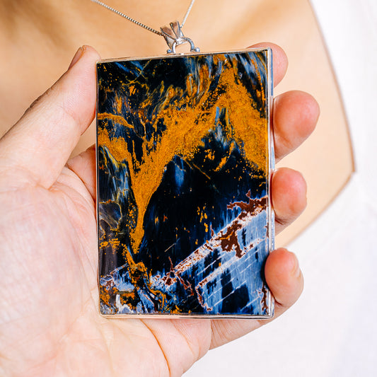Pietersite pendant close-up, rectangular stone with silver frame; blue, gold, and black patterns.