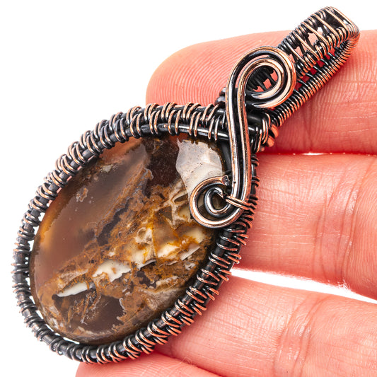 Oxidized copper wire-wrap pendant close-up: oval plume agate in coiled bezel with bail loop