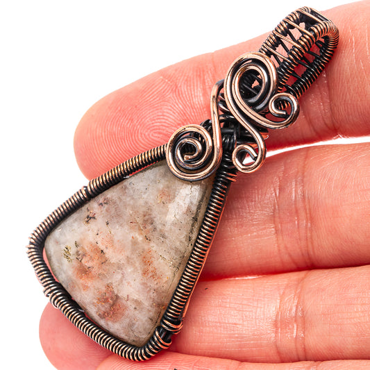 Oxidized copper wire-wrap pendant held between fingers, triangular stone with red-brown speckles.