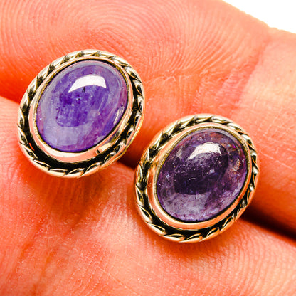 Oval tanzanite stud earrings on fingers, purple gemstone in gold-tone rope bezel.