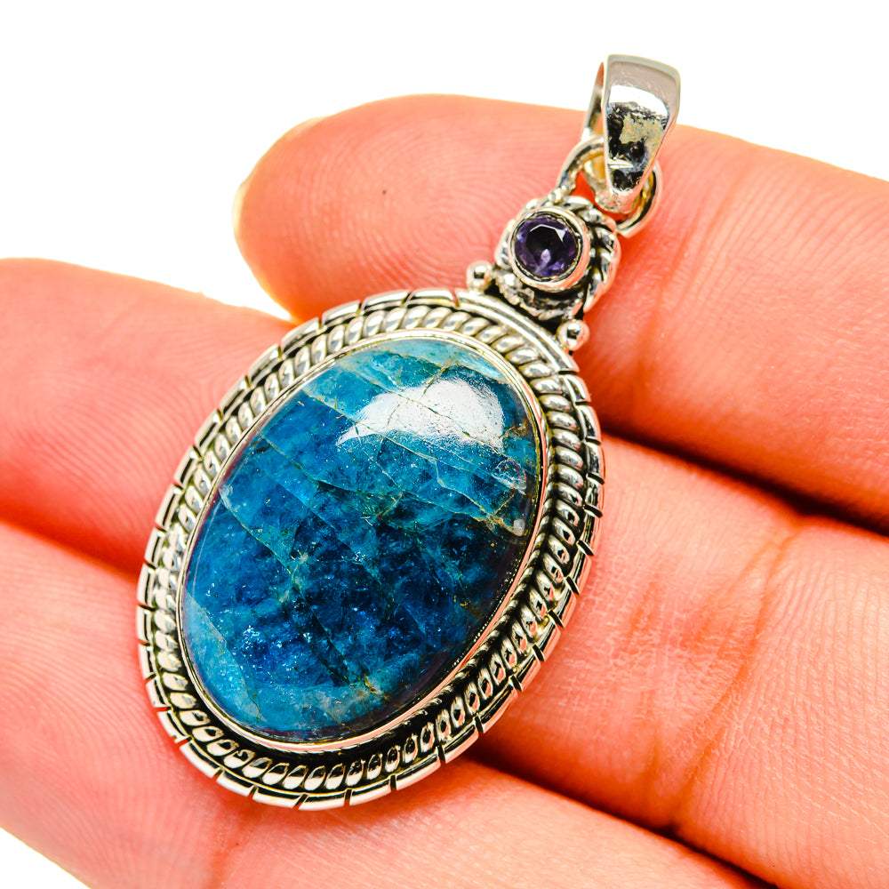 Oval blue gemstone pendant held on finger, silver setting with small purple top stone