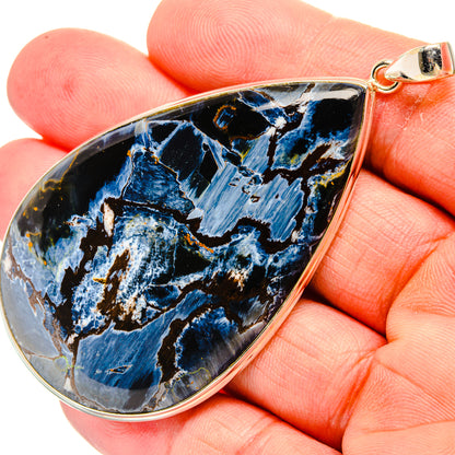 Pietersite teardrop pendant in silver-tone bezel with blue stone, held by hand.