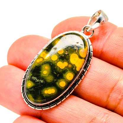 Oval ocean jasper pendant in hand, green stone with yellow spots, silver bail and frame.